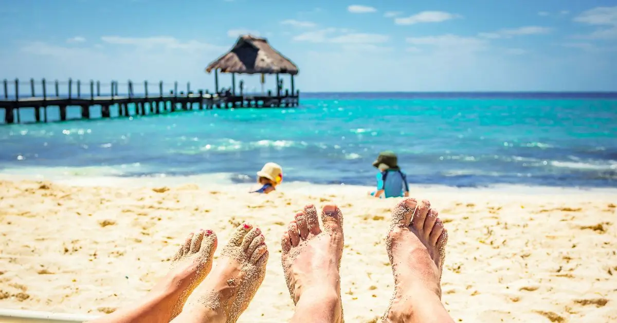 image captures a relaxing moment on Paradise Beach in Cozumel, Mexico. In the foreground, two pairs of sandy feet are stretched out on the beach, with a view of the turquoise sea and a pier with a thatched hut in the distance. Two children are playing in the sand, adding a playful element to the serene beach scene.