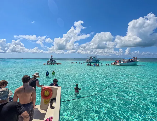 Tour boats anchored at Stingray City sandbar in Grand Cayman with groups of visitors standing in the clear turquoise water under a bright blue sky.