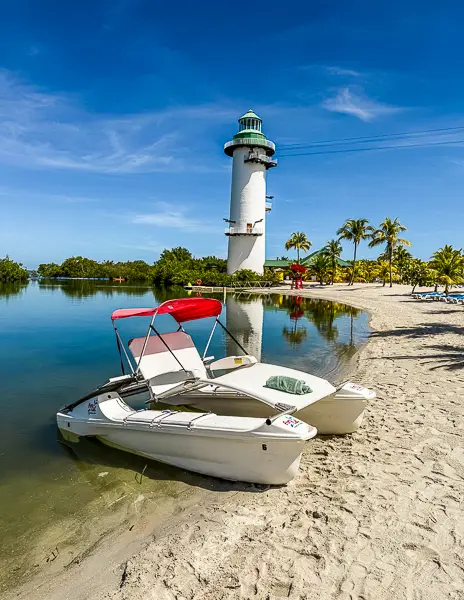 Paddle boats resting on the sandy shore beside the lighthouse at Harvest Caye, Belize, part of Norwegian Cruise Line’s private island, on a sunny day.