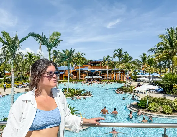 Cruiser (Kathy) overlooking the resort-style pool at Harvest Caye, Belize, part of Norwegian Cruise Line’s private island, with palm trees, loungers, and the LandShark Bar & Grill in the background.