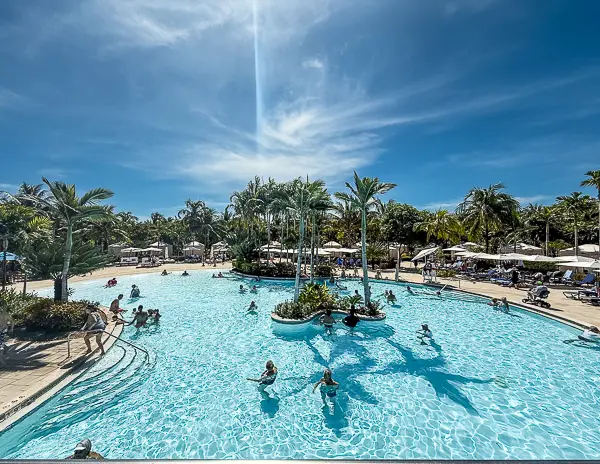 Guests enjoying the large resort pool at Harvest Caye, Belize, on Norwegian Cruise Line’s private island, surrounded by palm trees, loungers, and tropical landscaping under a bright blue sky.