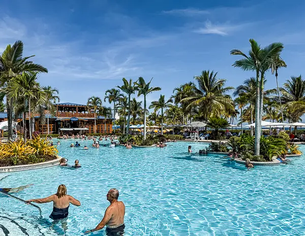 Guests enjoying the large resort-style pool at Harvest Caye, Belize, part of Norwegian Cruise Line’s private island, with palm trees, loungers, and the LandShark Bar & Grill in the background.