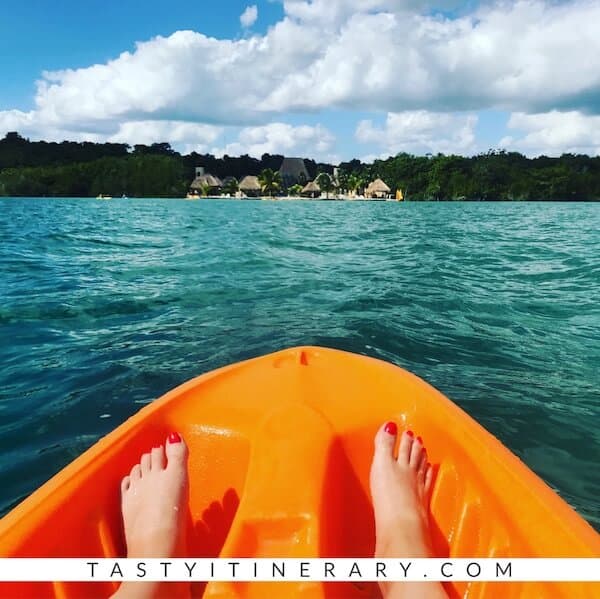 Tip of an orange kayak, pair of feet, pointing towards huts on a lagoon in Costa Maya