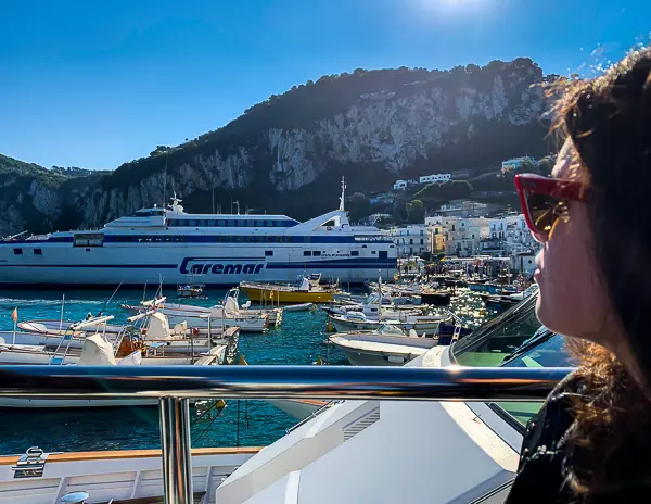 View of the Caremar ferry docked at Marina Grande in Capri, Italy, with small boats in the harbor and a woman (kathy) in sunglasses looking out from a railing in the foreground.