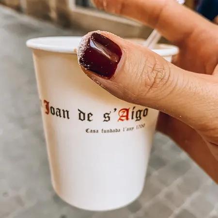 A hand holding a cup of café con leche from Can Joan de s’Aigo in Palma de Mallorca, with sugar-dusted fingers after enjoying an ensaimada.