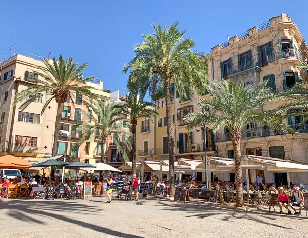 A lively plaza in Palma de Mallorca, Spain, with outdoor cafés, palm trees, and colorful historic buildings under a bright blue sky.
