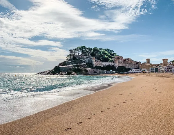 The shore to the left and sand to the right with foot prints leading up to castle ruines perched on a hill in Tossa del Mar