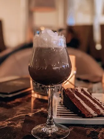 Specialty coffee drink served in a wine glass with whip cream and a slice of chocolate cake behind it.