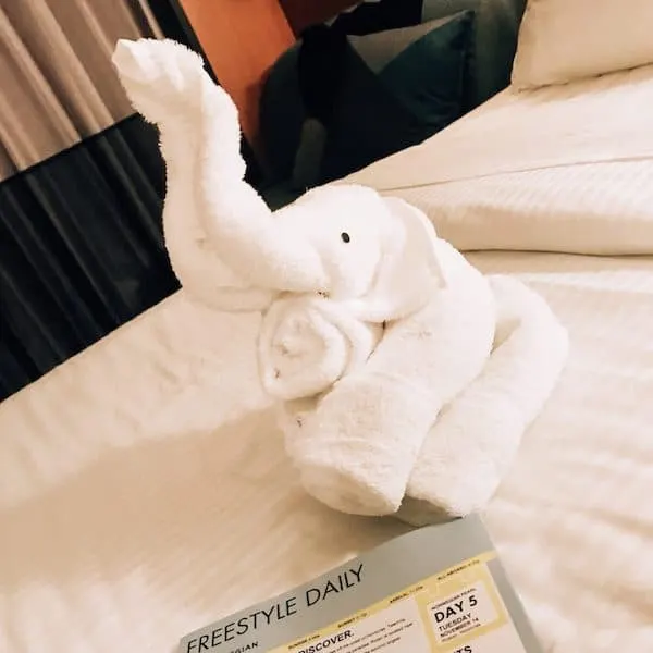 A towel elephant placed on a neatly made cruise cabin bed, accompanied by the Freestyle Daily itinerary, showcasing a charming detail from Norwegian Cruise Line's former turn-down service.