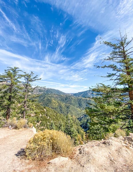 A scenic mountain view in San Bernardino National Forest near Big Bear Lake, with lush green trees, rugged terrain, and a bright blue sky with wispy clouds.
