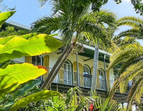 The outside of Ernest Hemingway's home in Key West, viewed through vibrant tropical foliage, highlighting the home's iconic yellow exterior and green shutters on the upper balcony.