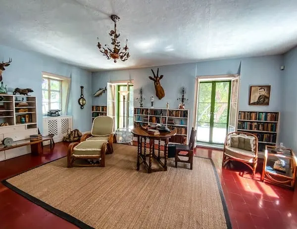 A spacious room that served as Ernest Hemingway's office, furnished with vintage chairs, a large rug, and walls adorned with hunting trophies and bookshelves filled with books.