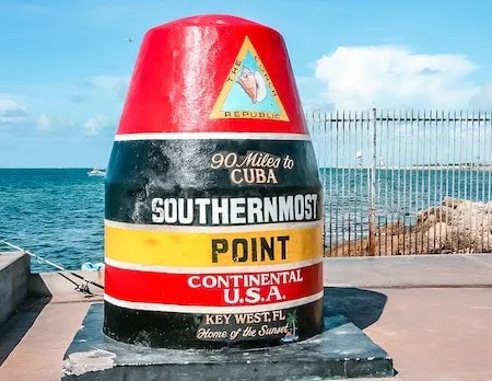 The Southernmost Point buoy in Key West, Florida, a colorful landmark indicating that it is just 90 miles to Cuba. The buoy is painted with vibrant bands of black, yellow, and red, and labeled "Southernmost Point Continental U.S.A., Key West, FL Home of the Sunset."
