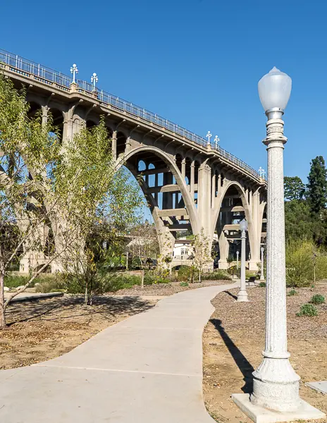 View of the Colorado Street Bridge from Desiderio Neighborhood Park in Pasadena, California, with a walking path and vintage-style lamp posts in the foreground.