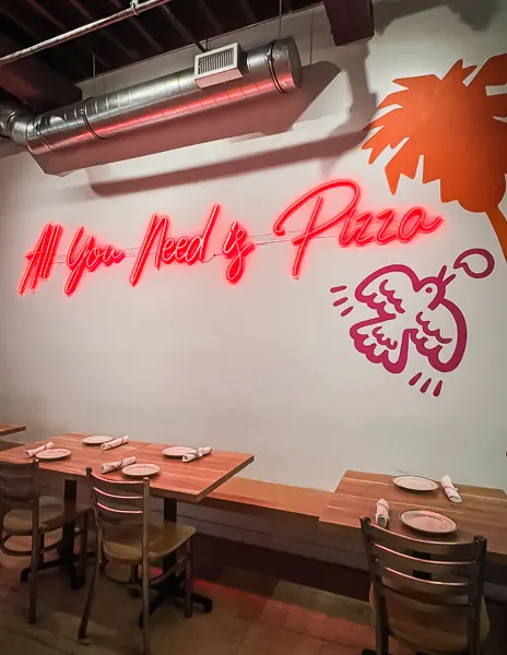 A neon sign on a wall stating "All You Need is Pizza" next to a stylized image of a pizza slice.