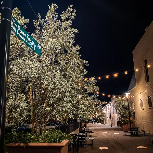 Nighttime view of Big Bang Theory Way in Pasadena, with streetlights illuminating the path and a street sign marking the location, surrounded by trees and festive lights.