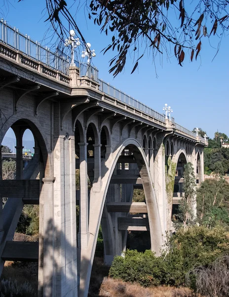 The Colorado Street Bridge in Pasadena, California, captured from below, showcasing its grand arches and architectural details surrounded by greenery.