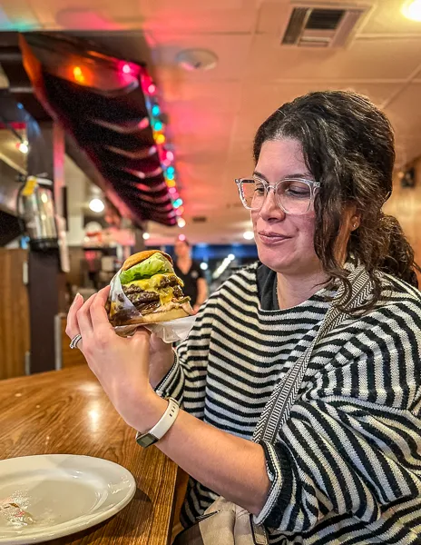 Kathy admiring a large, juicy burger she's about to eat at a diner with colorful string lights in the background.