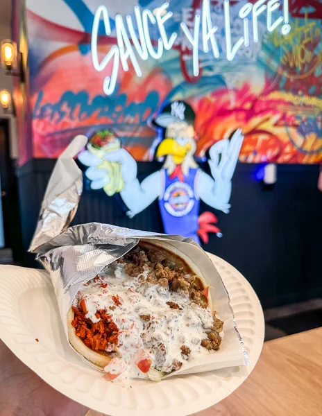 holding up a wrapped gyro with sauce on it and a mural of a chicken in the background