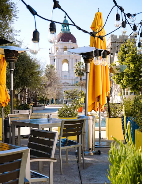 Outdoor seating area at California Pizza Kitchen in Pasadena, with bright yellow umbrellas, a clear view of Pasadena City Hall in the background, and decorative string lights.