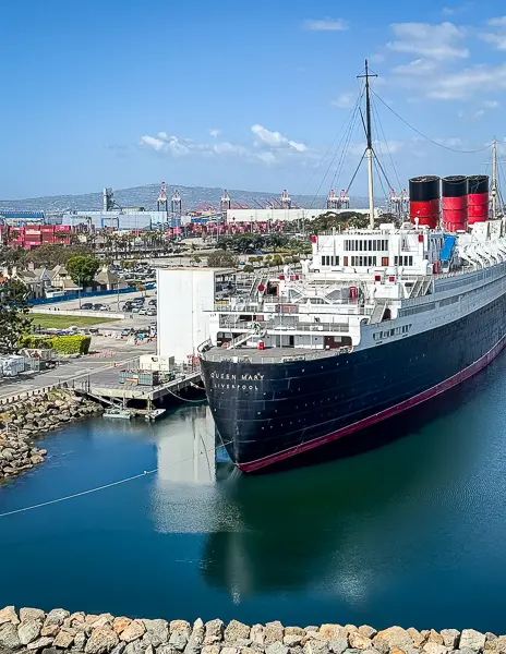 Historic ocean liner RMS Queen Mary docked in Long Beach, California, with its distinctive black hull, red funnels, and surrounding port facilities in the background.