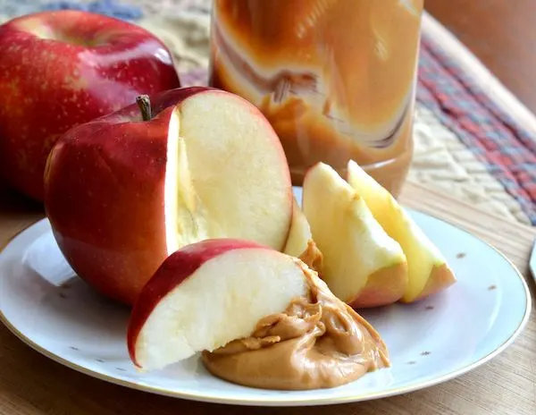 an apple with a few slices dipped in peanut butter adn the jar of peanut butter behind itr