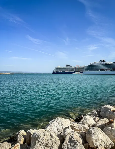 Celebrity Beyond and Viking Neptune cruise ships docked side by side at a Mediterranean port in October, viewed from a rocky shoreline with calm turquoise water and a clear blue sky.