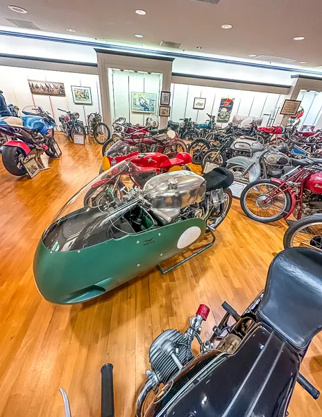 A vintage racing motorcycle with a sleek, green fairing, showcased in a spacious museum setting among other classic bikes.