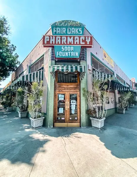 Exterior view of Fair Oaks Pharmacy & Soda Fountain in South Pasadena, featuring its vintage sign celebrating 100 years, with the storefront and potted plants lining the entrance.
