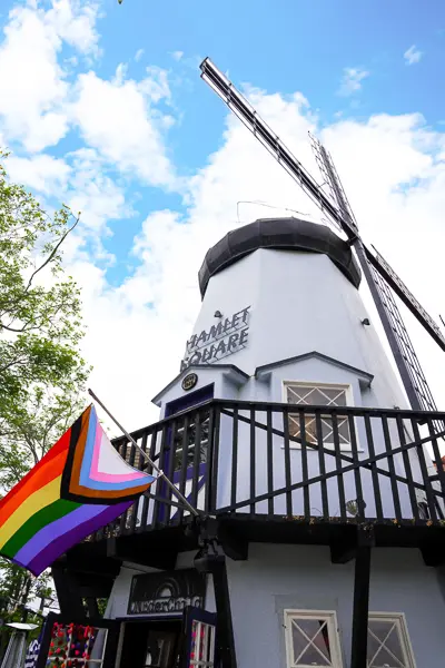 A windmill above Hamlet Square in Solvang, with a pride flag fluttering from a balcony against a backdrop of blue sky.