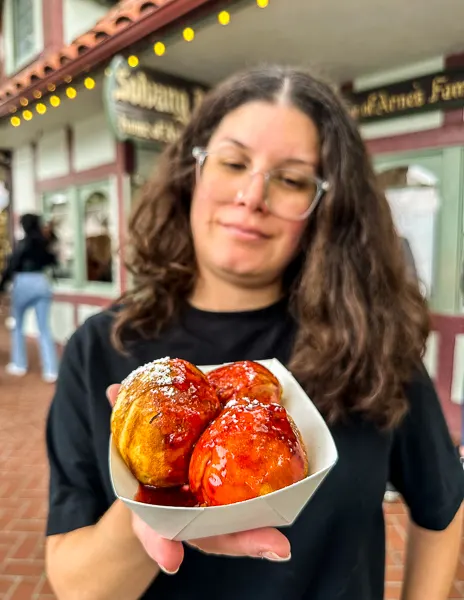 kathy holding a small container of fresh Aebelskivers, Danish round pancakes, dusted with powdered sugar and glazed with raspberry jam, with a blurred street scene in the background.
