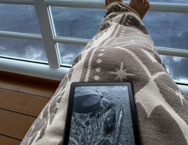 Reading a Kindle on a cruise balcony during a rainy sea day with ocean views in the background