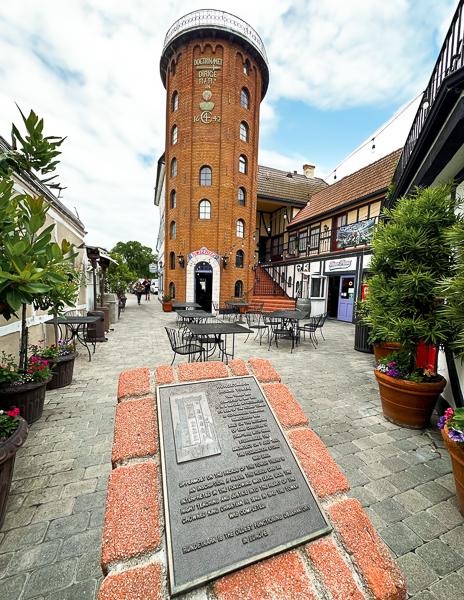 A pedestrian pathway leads to a tall, brick-built tower with decorative elements in Solvang, flanked by outdoor café seating and traditional Danish-style buildings.
