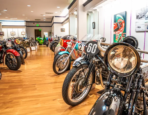A row of classic racing motorcycles with prominent number plates, lined up inside a well-lit vintage motorcycle museum.