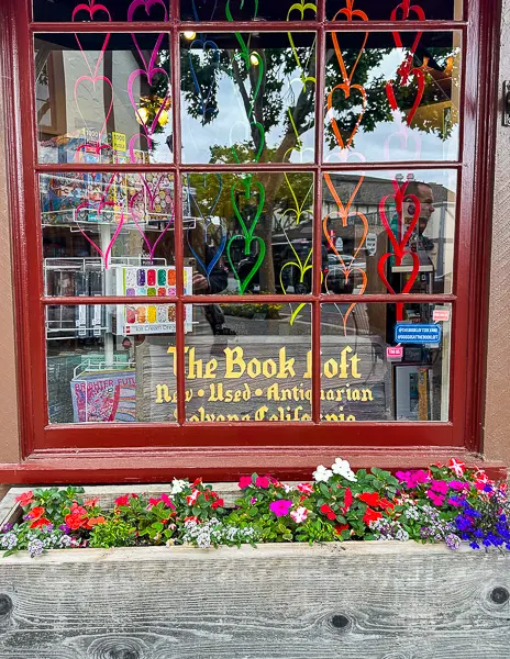 A cozy bookstore window with colorful heart decorations and a flower box filled with vibrant blooms and the book loft sign in Solvang, California