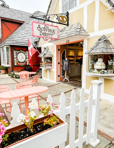 The exterior of The Solvang Bakery, a charming Danish-style shop with a pink sign, outdoor seating, and a white picket fence in Solvang, California.