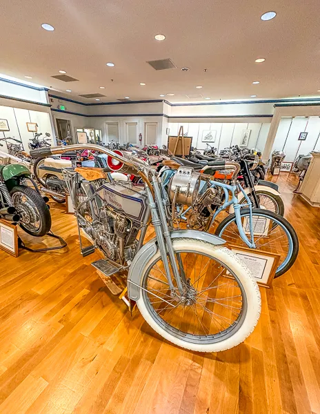 A collection of vintage motorcycles with unique designs displayed in an orderly row inside a museum with wooden flooring.