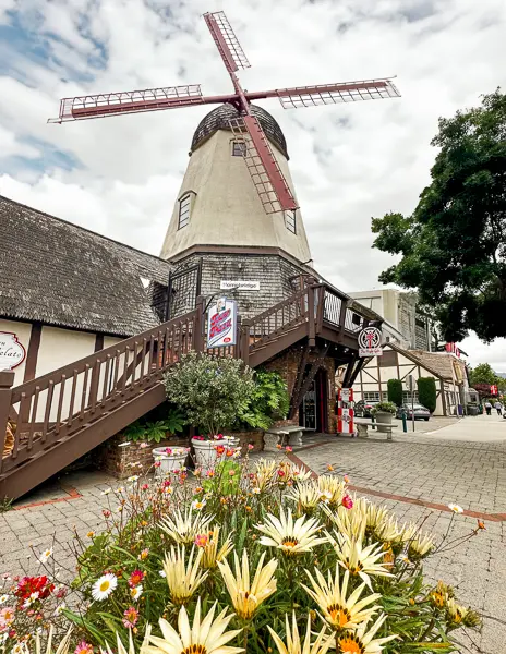 A windmill towers over a street in Solvang, with a flowerbed in the foreground adding a splash of color to the scene.