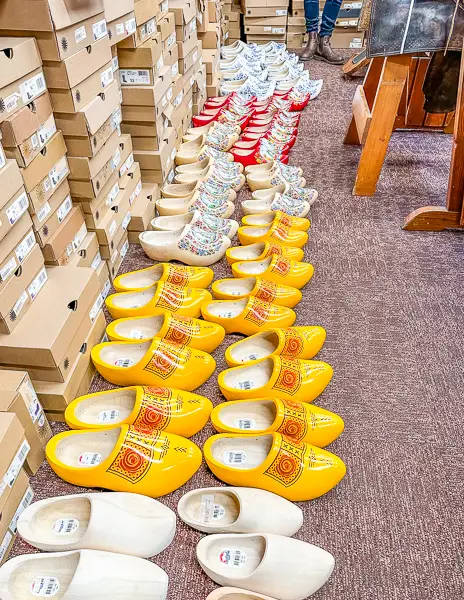 Rows of colorful wooden clogs, predominantly yellow and decorated with intricate patterns, lined up inside a shoe store alongside boxed merchandise.