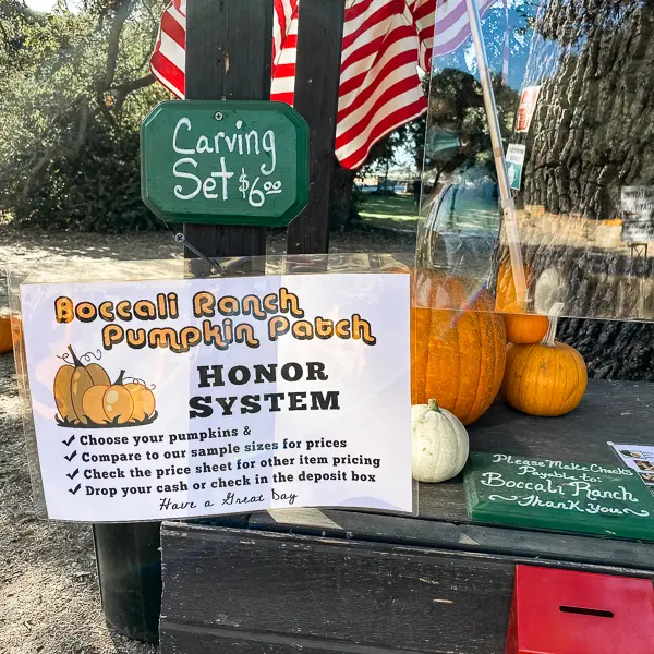 A sign at Boccali Ranch Pumpkin Patch in Ojai explaining the honor system for purchasing pumpkins, placed on a small wooden stand with pumpkins and an American flag in the background.
