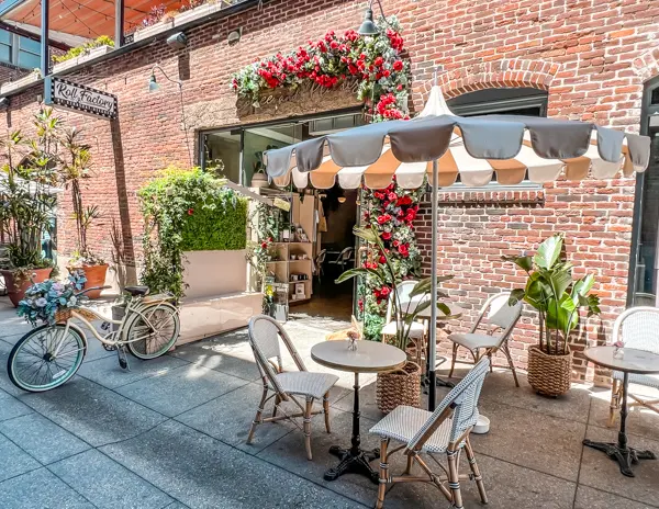 Charming outdoor seating area at Coffee and Plants in Old Pasadena, featuring bistro tables, a vintage bicycle, lush greenery, and red floral decor against a brick wall.