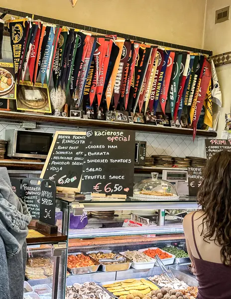 the counter at Pino's Sandwiches, with a display of fresh ingredients and a colorful array of regional flags hanging overhead, showcasing the day's special focaccia sandwich