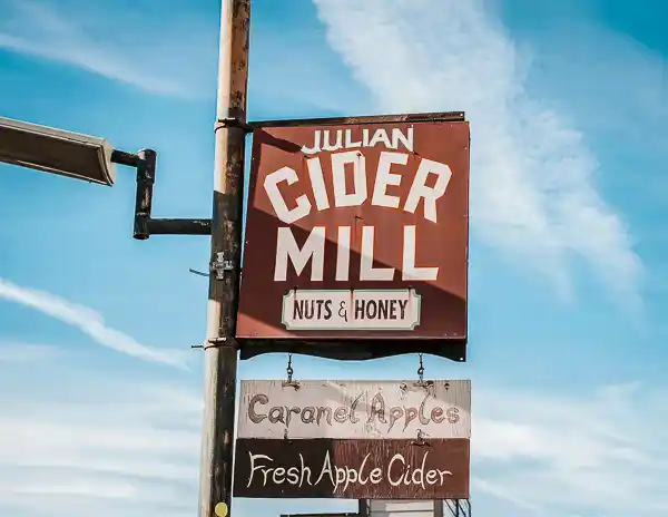 A rustic sign for Julian Cider Mill reading “Cider Mill, Nuts & Honey” with a smaller sign below advertising “Caramel Apples” and “Fresh Apple Cider,” set against a blue sky.