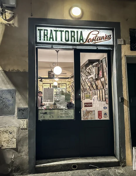 The entrance to Trattoria Sostanza in Florence, Italy, a traditional eatery with a glowing sign above the door and a glimpse of the cozy interior.