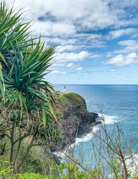 "Kilauea Lighthouse perched on a rocky cliff overlooking the blue ocean, surrounded by lush greenery and under a partly cloudy sky in Kauai, Hawaii.