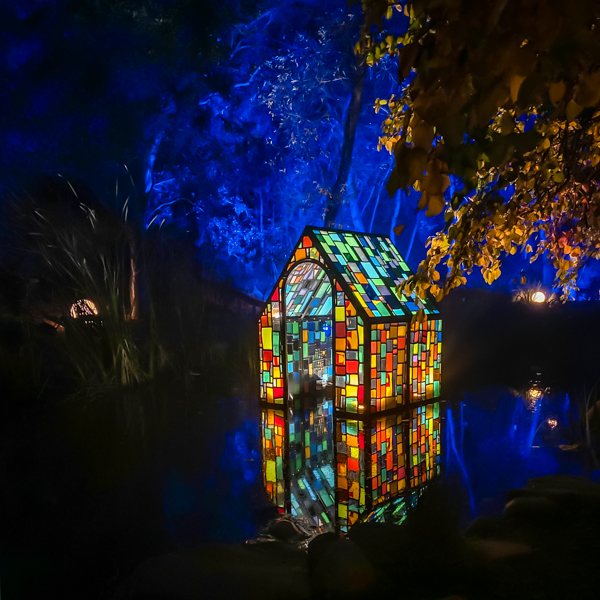 A vibrant, illuminated structure resembling a colorful stained-glass house reflecting on a pond, part of the Enchanted event at Descanso Gardens.