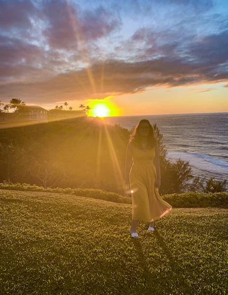 Person (kathy) in a yellow dress standing on a grassy hill overlooking a stunning sunset over the ocean in Princeville, Kauai, with soft sunlight and clouds in the sky.