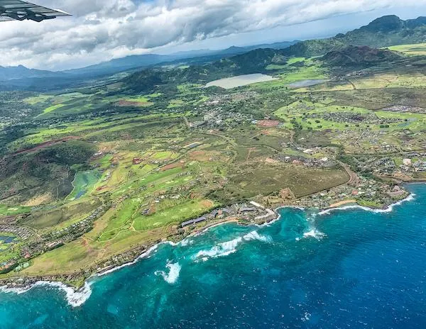 Expansive aerial view of Kauai's southern coast, featuring rolling green fields, a vibrant coastline with turquoise waters, and scattered buildings.