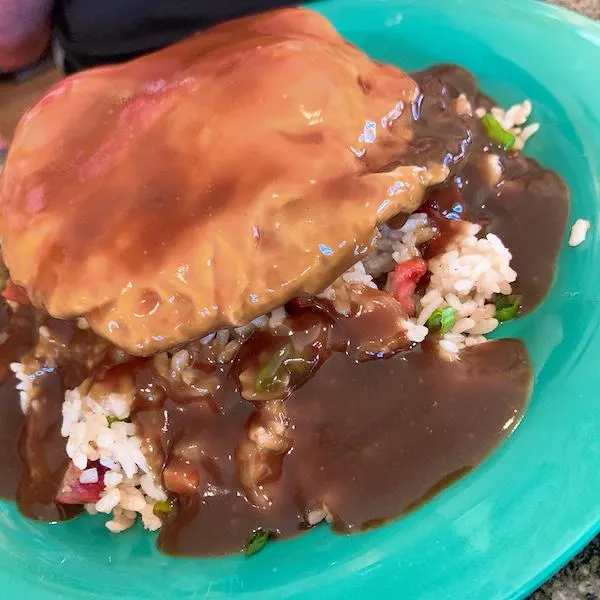A plate of Hawaiian Loco Moco featuring a hamburger patty, fried egg, and savory brown gravy served over rice with colorful vegetables on a green dish.