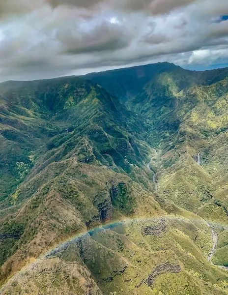 Spectacular aerial view of Kauai's rugged mountains with deep valleys, waterfalls, and a vibrant rainbow arching over the landscape.